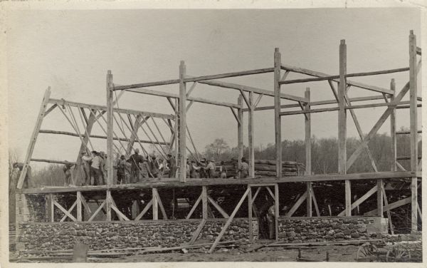 A large group of men are using log poles to pull the side of a new timber framed barn up to meet the roof line. 
