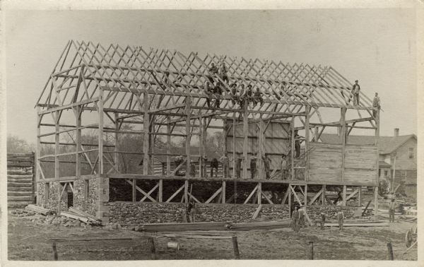 A large group of men pause briefly in their work of raising this large timber framed barn.