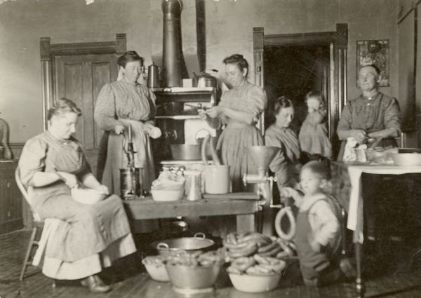 Three generations of the Fiebelkorn family make sausage in the family farm kitchen. On the table is a sausage stuffing implement, a meat grinder, and a sausage crock from which one woman is tying up sausage. Each person appears to have a task, such as mixing spices into the ground meat. One child is holding a ring bologna.