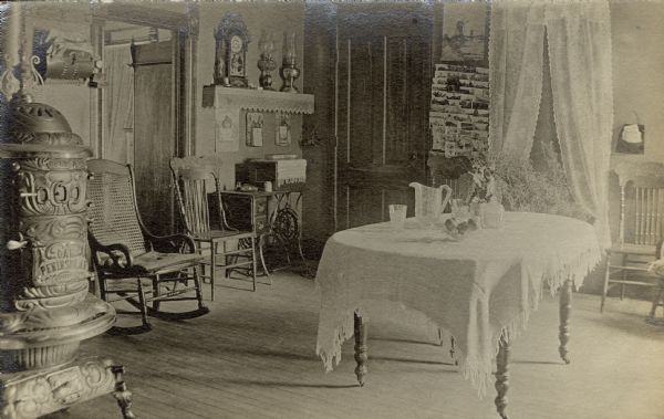 Interior of William Fiebelkorn's home. Next to a window with lace curtains at the window is a rack with a display of photographs or postcards. There is a treadle sewing machine in one corner, a rocking chair, and a dining table in the center of the room set with a pitcher and glasses. A wood burning stove is on the left in the foreground.
