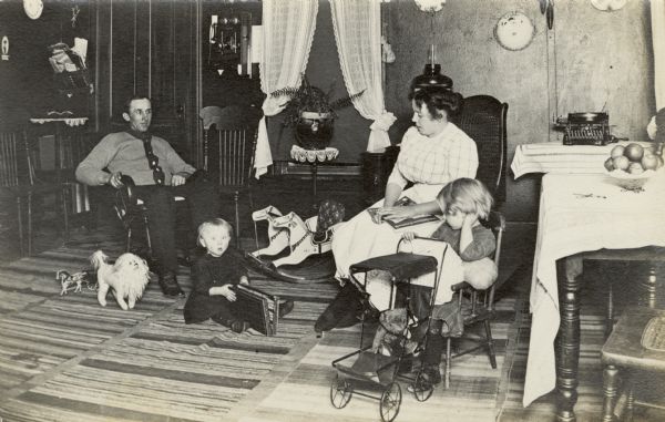 William Fiebelkorn's family sit together in a sitting room. The two children pose with toys. There is a stuffed animal near William's feet. Anna Fiebelkorn is holding a magazine in her lap. There is a typewriter on a table on the right, and on another table is a bowl of fruit and a pair of scissors. A telephone is on the wall by the door in the center.