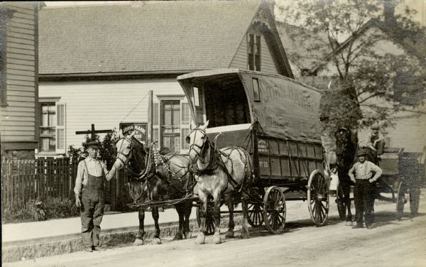 John Hahn stands in the street near his horse-drawn cart. The side of the cart says 'John Hahn Piano & Furniture Moving'. The cart, John Hahn, and the cart driver are standing in front of John Hahn's home at 320 Lapham Street, Milwaukee. The sign in front of the house, partially obscured by the horse, says John Hahn City Express and Furniture Moving. Behind the moving wagon is a second horse-drawn vehicle with a driver.