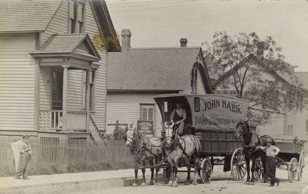 View from the street of John Hahn standing on the right with a horse hitched to a passenger wagon. In the center is the furniture moving wagon he owns, in which another man sits holding the reins of two horses. The two wagons are parked outside John Hahn's home at 320 Lapham Street. A small boy with a kite, possibly John Hahn's son, is standing outside the house near the fence. There is a another child, a toddler, sitting on the porch.