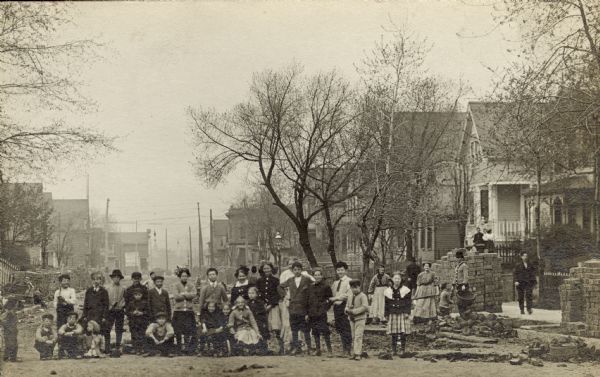 Group portrait of neighborhood children and adults gathered on a street in the S. Sixth Street neighborhood of Walker's Point that is being re-paved. Many of them are smiling broadly. Men and women are holding construction implements on the right side of the street near piles of bricks.