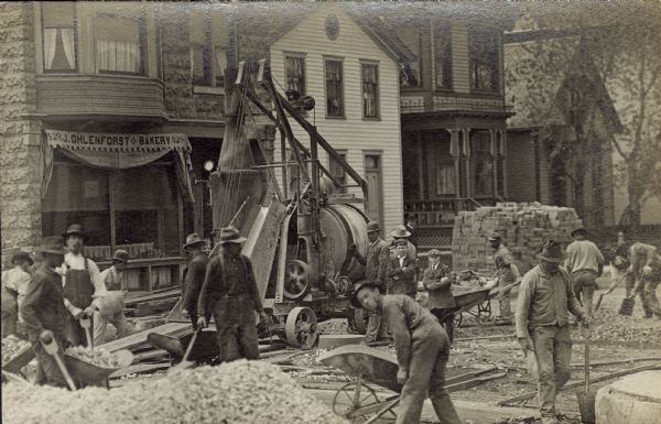 Traffic to the J. Ohlenforst Bakery at 529 S. Sixth Street, in the Walker's Point neighborhood, is impeded by street construction. There are piles of gravel and bricks waiting to be used. Many men with wheelbarrows and shovels are working.