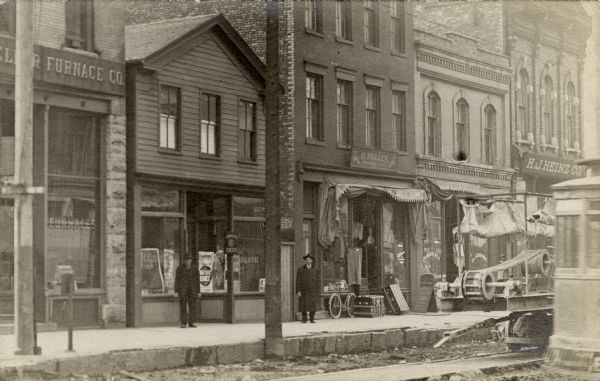 Street view in the S. Sixth Street neighborhood of Walker's Point. The storefronts include the H.J. Heinz Co.; H. Mullen, a clothing and shoe store; J. Gerstner, Tailor; and a furnace company. There are men standing outside two of the businesses. The street level is below the sidewalk and is unfinished. On the right in the foreground is a streetcar; next to it on parallel tracks is a construction machine.