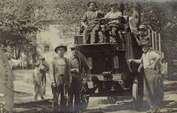 John Hahn is leaning on a motorized moving van in the S. Sixth Street neighborhood of Walker's Point. At the steering wheel is a man, with another seated next to him. A man behind John Hahn is standing on the side of the van. Two men in overalls stand in the street. Behind them on the sidewalk are two children.