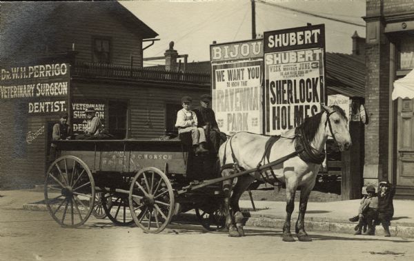 A horse-drawn cart, marked B.C. Horsch, is waiting near Dr. W.H. Perrigo's veterinary and dental business in the S. Sixth Street neighborhood in Milwaukee. Behind the horse are commercial buildings. Two billboards advertize nearby theaters, the Bijou and the Shubert. The Shubert will begin showing Sherlock Holmes for a week beginning Monday June 3, 1907. In the front of the Horsch cart are a boy and a man, and two more boys sit on the back. There are two children sitting on the curb in front of the cart. The Shubert Theatre site is now occupied by the Pioneer Building.