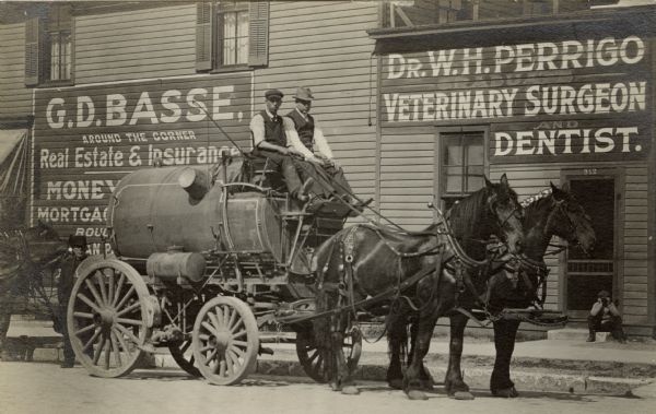 Two men sit on top of a water tank wagon which is pulled by two horses. They are posing in front of the W.H. Perrigo Veterinary Surgeon and Dentist office on the right. There is a sign painted on the building for the G.D. Basse Real Estate and Insurance office, which is around the corner.