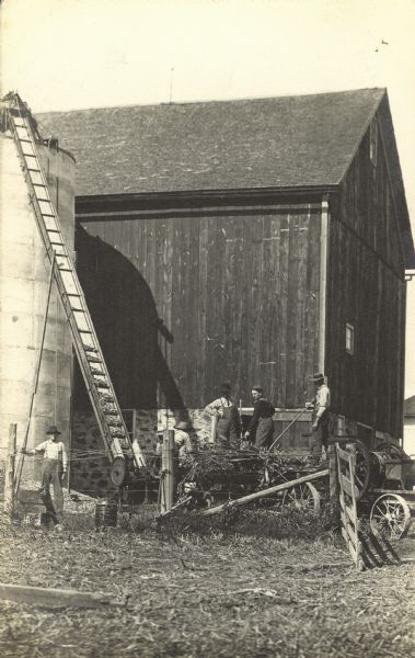 View across barnyard towards five men on the William Fiebelkorn farm working with a wagon and conveyor belt to load hay into a silo. The barn has a stone foundation. There is a barbed wire fence with an open gate in the foreground.