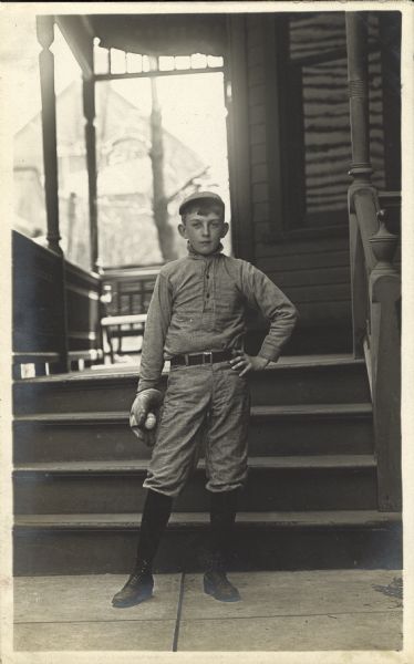 A young baseball player poses outside the porch of his S. Sixth St. Walker's Point neighborhood home. He is wearing a baseball uniform with cap, and is holding a catcher's mitt and ball.