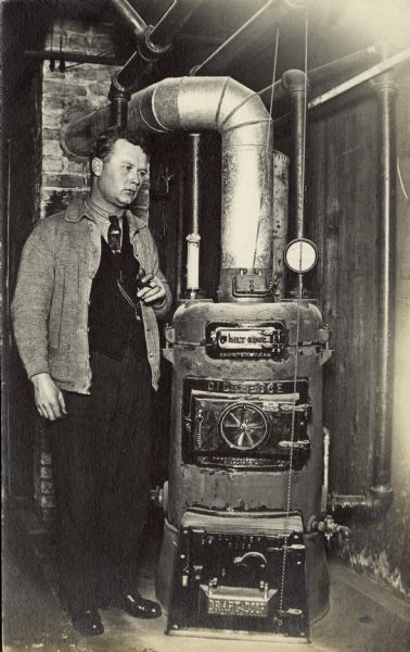 A man holding a cigar stands near a Gilt Edge Liberty furnace. The R.J. Schwab and Sons Co. in Milwaukee sold Liberty and Pipeless furnaces for over fifty years. The man is wearing a necktie, sweater, and vest and wears a watch on a chain.