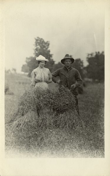 A young woman and man pose behind a hayrick in a farm field. They both wear hats.