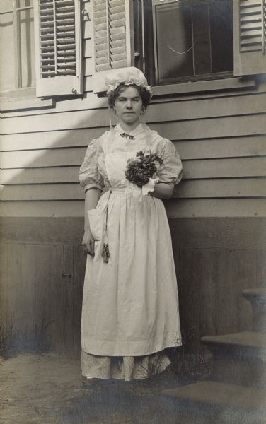 A young woman dressed as a maid in a long apron and wearing a mob cap, stands outdoors against the side of a house. She has a set of keys hanging from her waist on a lanyard, and she is holding a bouquet of roses.