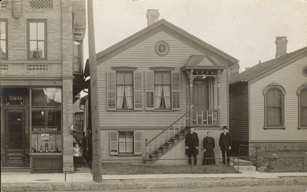 View from the street of three people standing in front of a small house. There is a small Oeil-de-boeuf window in the upper story. The windows have shutters and lace curtains and decorative wooden window heads, and the porch has spindled columns. A young boy and an older boy stand in the alley on the left. There is a brick building with a sign for the "First Ave. Exchange" on the left with an address of 520. On the right is another small house.<p>The postcard text is in German. It is addressed to Mr. William Fiebelkorn, Cascade, Sheboygan Co. Wisconsin.
