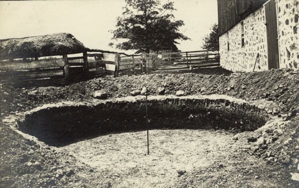 The hole for the new silo on the William Fiebelkorn farm. A pole marks the center. There is a barn on the left with a stone foundation. In the background are fences and enclosures.
