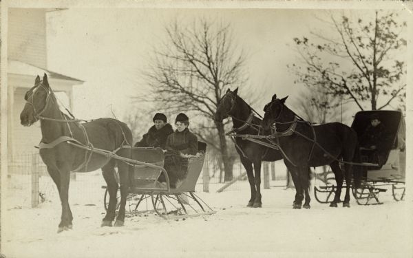 Two horse-drawn sleighs are parked in the snow in front of a house. In the first one on the left, William Fiebelkorn Sr. and his wife Fredericke (Krause) sit with furs on their laps and dressed in warm coats and hats. Their sleigh is hitched to a horse. The second sleigh has two horses and a single man sitting in it.