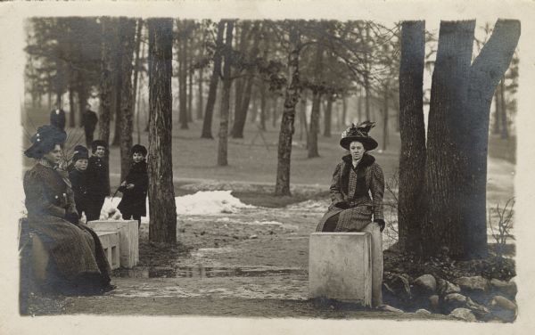 Two women sit pensively on stone benches on either side of a muddy gravel path. The one on the left is Elsie. Near her in the background are four children. In the far background, two men stand among the trees.<p>The postcard is addressed to Mrs. Wm (Anna Unglaube) Fiebelkorn in Cascade, Wisconsin. The text is: "This will give you a little reminder of how I look until next summer." It is signed "Elsie K."