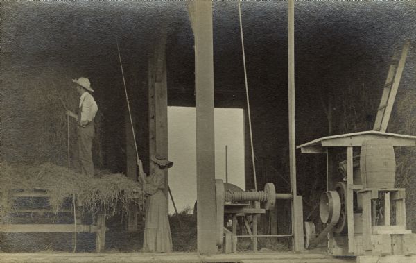 Two people are using a winch and pulley system to move hay into the hay loft. The woman is wearing a long dress and a ruffled hat. The man is wearing overalls and a hat and is standing on the wagon.