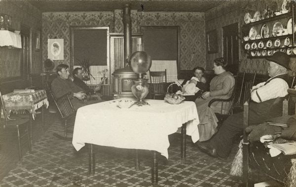 William Fiebelkorn's family gathered in a living room. William Sr. is in a rocking chair with his hands clasped. His brother August Fiebelkorn is reading a magazine. William's wife Fredericke (Krause) is holding a small baby. The older couple is perhaps Fredericke's parents. The room has a stove, a phonograph and a basket of fruit on a table with a lamp. There is a pile of knitting in the right foreground.