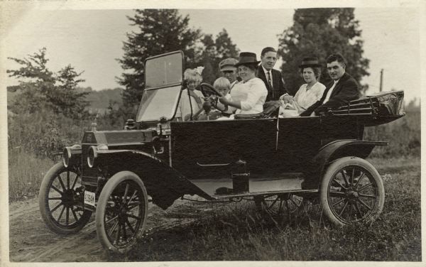 A smiling Anna Fiebelkorn is driving a touring car. William Fiebelkorn is next to her with two children in front of him. In the back seat are a woman with a baby and a man. Standing outside the car, perhaps on the running board, is another man.