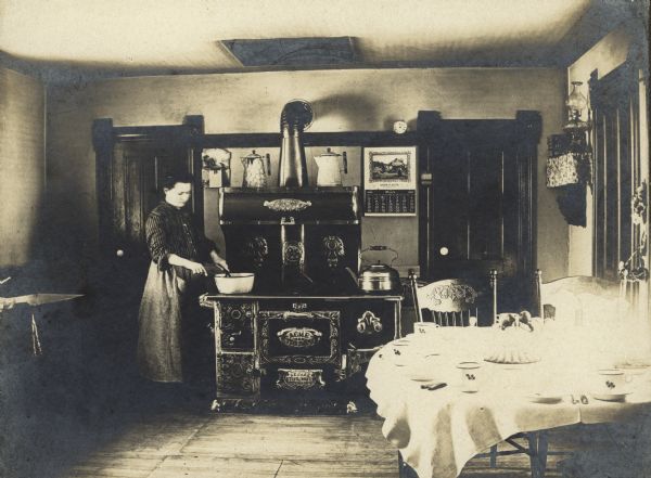 A woman is stirring a pot on what looks like a new stove. It is a gleaming Acme, from Newark Stove Works. There is a warming closet in the top half. The calendar on the wall shows that it is March 1905. The table is set for a meal.