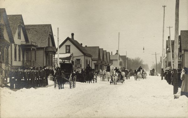 View down a snowy street. Men and women, one with a baby, are bundled up against the snowy winter weather, and are lining both sides of the street. Carriages are pulled up in front of a house, and more are coming down the street. The hearse is pulled by two white horses wearing fringed netting. On the far left a group of uniformed policemen stands on the sidewalk.
