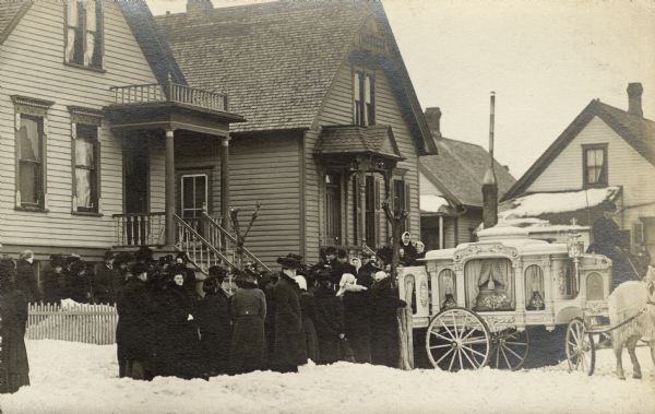 An elaborate funeral hearse is pulled up to a house with the back doors open, waiting to be loaded with the casket. There are many men and women bundled up in winter clothes waiting outside the house. Snow is piled in the yard and along the street. The hearse is pulled by two white horses wearing fringed netting.