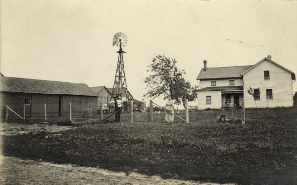 William Fiebelkorn and his wife Anna stand at a fence on their farm. William is holding a toddler. The house, the barn, several outbuildings and a windmill are in the background.