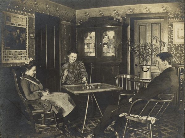 Anna Fiebelkorn sits at a table playing carrom with her husband William. One of her sisters is watching while sitting in a rocking chair on the left.