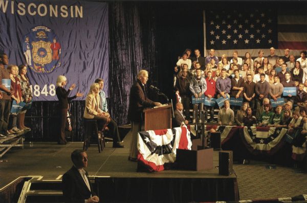 Vice President Joe Biden addresses the crowd at a 2012 Democratic Election rally held at the University of Wisconsin-La Crosse. He is standing at a podium on a stage, and behind him his wife, Dr. Jill Biden, sits on a stool. An enormous United States flag hangs on the right and a State of Wisconsin flag is on the left. Students are seated on the right and standing on the left, holding Obama-Biden signs. Patriotic bunting decorates the stage and the front of the bleachers.