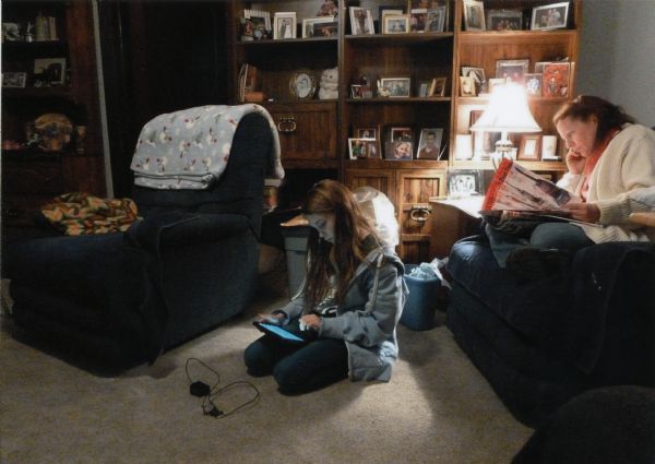 Mary (Kate) Marcou Temp is sitting on the sofa reading a newspaper while her daughter Kate Temp is kneeling on the floor viewing her electronic reader on Thanksgiving Day. Mary is the photographer’s sister. In the background is a bookcase filled with framed photographs. A quilt is hanging over the recliner on the left.