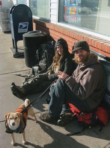 A young wandering couple, dressed warmly, sits on the sidewalk with their dog in front of a Kwik Trip store on Lang Drive. Their backpacks are resting beside them. The dog, wearing his own pack, is standing and peering up at the photographer. Signs are in the window behind them, and a trash can, mailbox and propane cylinder rack are along the sidewalk in the background.