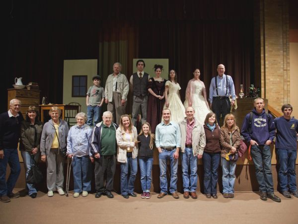 Group portrait of the cast, crew and extended family of David Marcou for the Pulitzer-nominated play "Remembering Davy Crockett," at the end of the world debut night at Aquinas High School. David Marcou, who wrote the play, is wearing a blue shirt with black suspenders, and is sitting at the far right in the back row. In the background are sets and props.