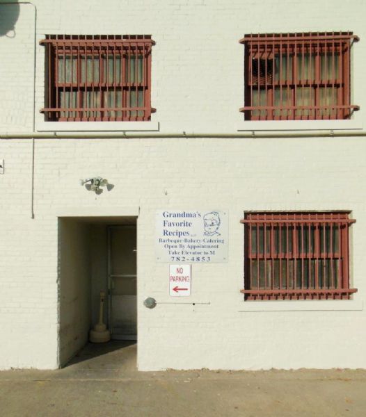 Sign mounted on a painted brick wall next to a doorway in a downtown alley. Features a head and shoulders caricature of a woman wearing glasses perched on her nose, and reads, "Grandma's Favorite Recipes LLC, Barbeque-Bakery-Catering." The sign also gives directions and a phone number. Above and to the right are three windows with red metal grills.