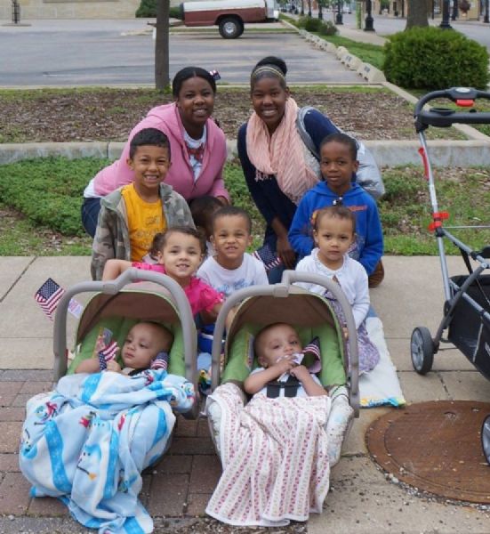 An African-American family poses on the sidewalk for a group portrait, while waiting at the corner of 6th & King Streets, for the Memorial Day Parade. The babies are in infant carriers and a stroller can be seen on the right. The children are holding small American flags.