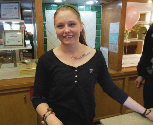 The hostess at a Perkins Restaurant smiling for a candid portrait. She has a “Time Is Precious” tattoo below her left collarbone, and is wearing a scoop-necked black shirt, a headband in her hair, earrings, a necklace, and bracelets on her wrists. The photographer can be partially seen in the background mirror.