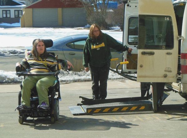 Crystal (Courtney) is assisted by a lift for her wheelchair into a van at Becker Plaza. The lift operator is wearing a Green Bay Packers sweatshirt. Building and snow can be seen in the background.