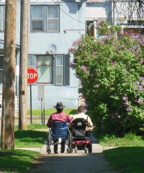 A married couple, Carrie and Prentice Vaughn, riding together into the distance in their wheelchairs on the sidewalk on 7th Street. On the right is a large lilac bush, on the left are a stop sign and power poles. In the background are buildings.