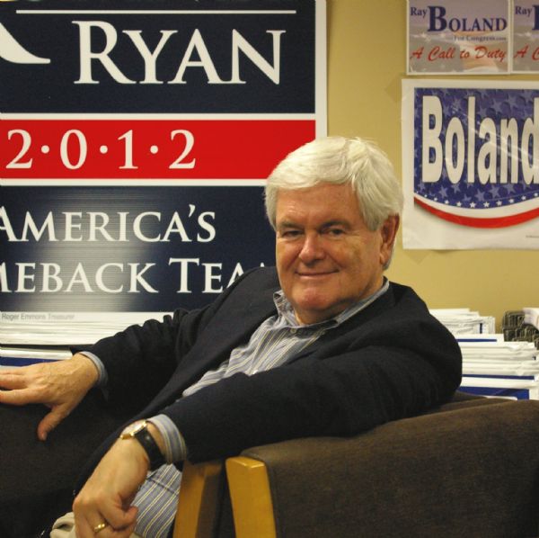 Former United States Speaker of the House Newt Gingrich, seated and smiling for the camera at a 2012 Election Rally. The rally was held at the La Crosse County Republican Headquarters. In the background, Romney-Ryan and Ray Boland campaign signs cover the wall.