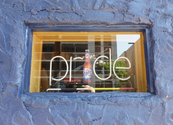 A neon sign with a bottle of Miller Lite for the "i" spells “Pride” in the window of the "Chances R" gay bar on Jay Street. The word "Pride" is underlined with a neon rainbow. The window is framed in wood and surrounded by stucco painted blue.