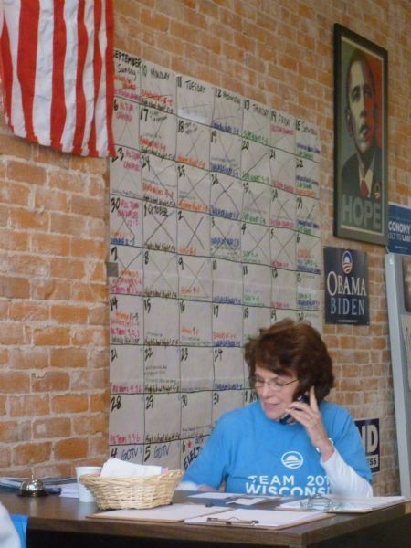 Mrs. Curt Reithel, a La Crosse County Democratic Headquarters worker, on the phone during the 2012 Election run-up. She is seated at a desk. A brick wall in the background is covered with a large calendar, American Flag, Hope and Obama-Biden posters.