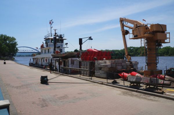 United States Coast Guard boat and a dredging barge at the Riverside Park, Levee Dock, a United States port facility on the Mississippi River. A brick walkway runs along the shore. In the background are the river and far shoreline with trees. On the left, in the distance, the U.S. Highway 14, 61 and State Highway 16 bridge can be seen.