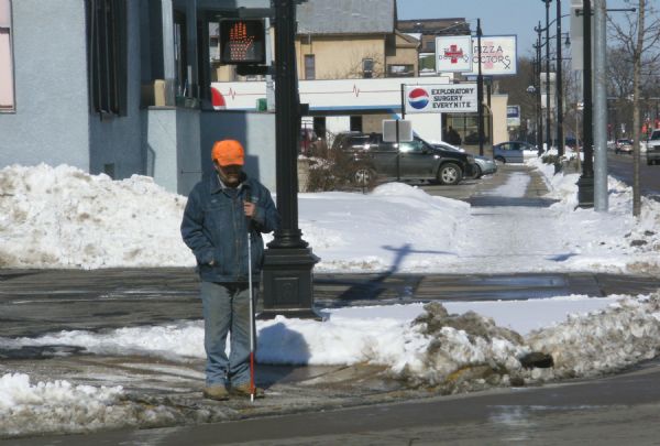 A blind man with a white cane waits on the corner of 7th & Cass Streets. He is wearing jeans, a denim jacket, blaze orange hat and boots. Snow is piled on the curb. In the background are a Pizza Doctors restaurant, other buildings, automobiles and streetlights.