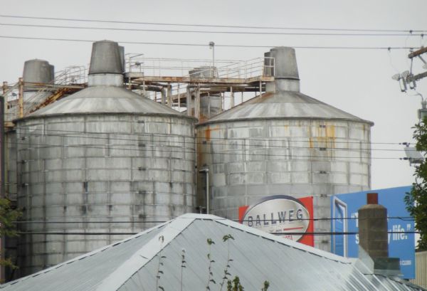 Large round grain elevators near 3rd Street. On the right are two billboards advertising "Ballweg Midwest Toyota" and "Boys & Girls Clubs." In the foreground is a metal roof and brick chimney.