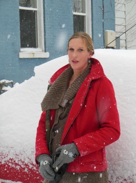 Beth, a neighbor of the photographer, smoking during a snowstorm on 7th Street. She is wearing a brown sweater with a shawl collar, an unzipped red coat, and grey gloves embroidered with flowers. She is standing in front of a snow covered automobile holding a snow shovel. In the background is a building painted blue with white trimmed windows.