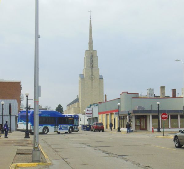View down street towards men at work on a boarded-up building on a street corner. A city bus enters the street on the left. In the background is the Cathedral of St. Joseph the Workman on 6th Street.