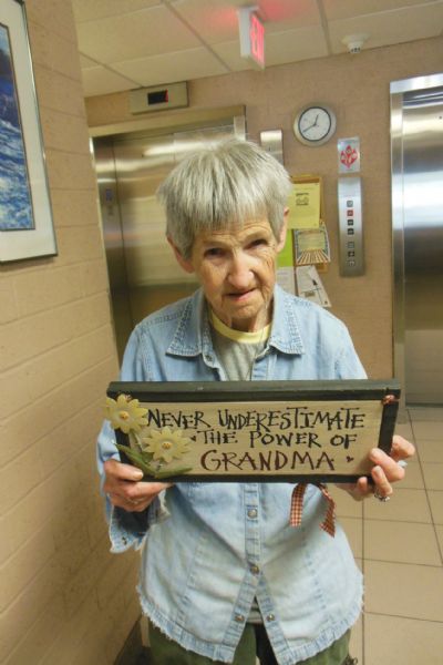 Lois Wason displaying her hand-painted saying about grandmothers, with flowers, on a board at Becker Plaza. She is wearing a light blue work shirt over a t-shirt. In the background are two elevators.