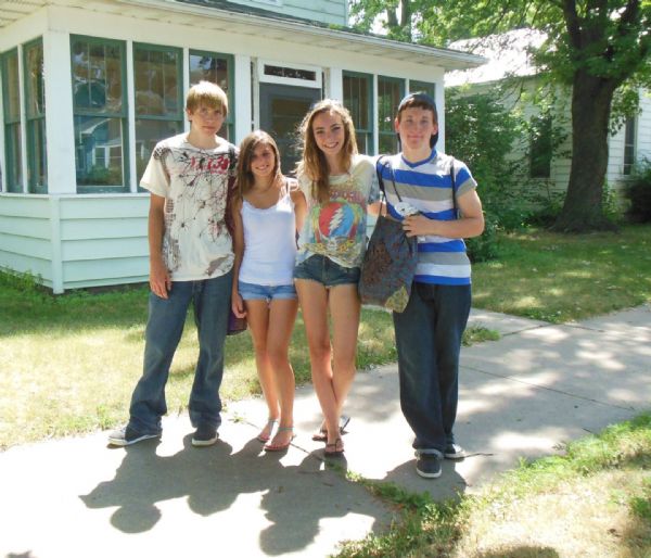 Four teens, two girls and two boys, pose together on the sidewalk on 7th Street. They are dressed casually, the girls in shorts and sandals and the boys in blue jeans and sneakers. One girl wears a white tank top and the others colorful t-shirts. The boy on the right is carrying a cloth tote bag. In the background is a porch with glass windows surrounded by a lawn. On the right is another house and a tree.