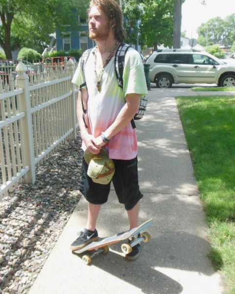 Sean Murphy, a professional skateboarder and snowboarder, poses on the sidewalk with his skateboard. He is gazing to the left while holding his baseball cap in his hands, and wears a tie-dyed t-shirt. On his back is a plaid backpack. He has ear buds in his ears. On the left is a white picket fence, on the right a car is parked at the curb. In the background are trees and a building.